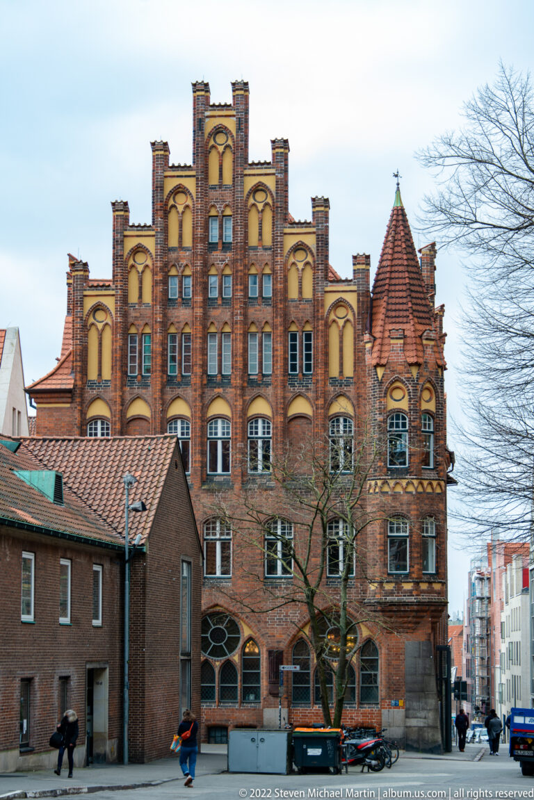 Large red-brick building with stepped gable, recessed arches, rows of windows, and a corner turret.