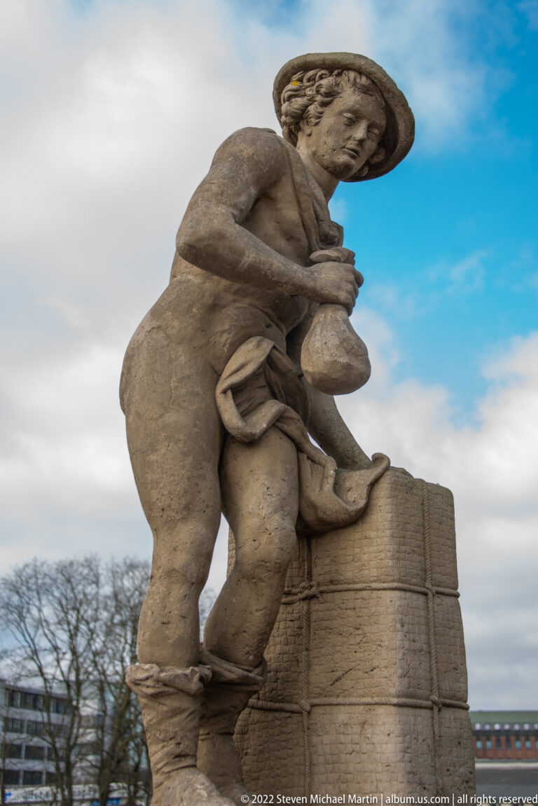 Stone male figure in a broad hat bending beside a masonry block while holding a rounded vessel.