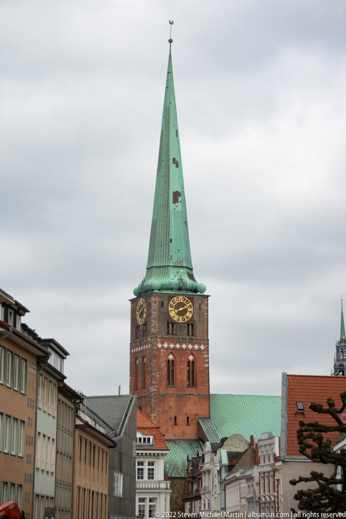 Red-brick church tower with large clock faces and a steep green copper spire above city rooftops.