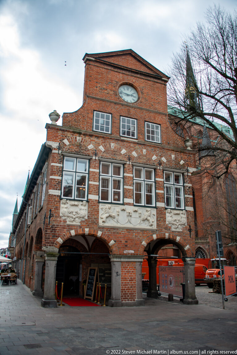 Low historic brick building with broad ground-floor arches, white-framed upper windows, and service equipment outside.