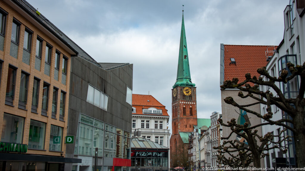 Green-spired brick church tower with clock face at the end of a narrow street lined with shops and pollarded trees.