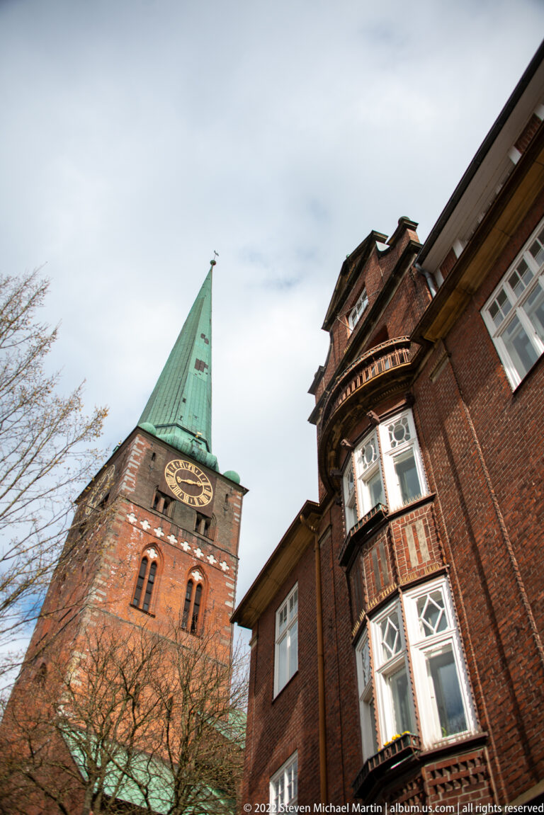 Green-spired brick church tower behind a historic brick house with a projecting polygonal bay window.