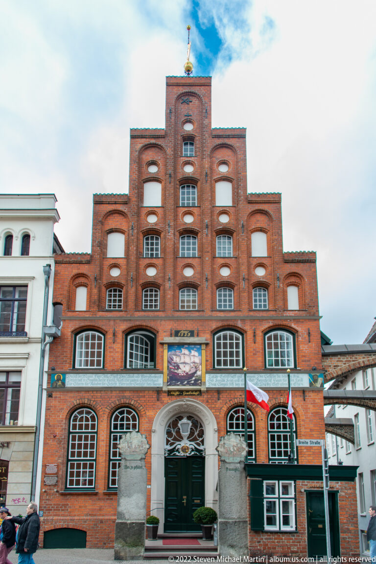Red-brick stepped-gable building with rows of arched openings, a carved stone entrance, and broad ground-floor windows.