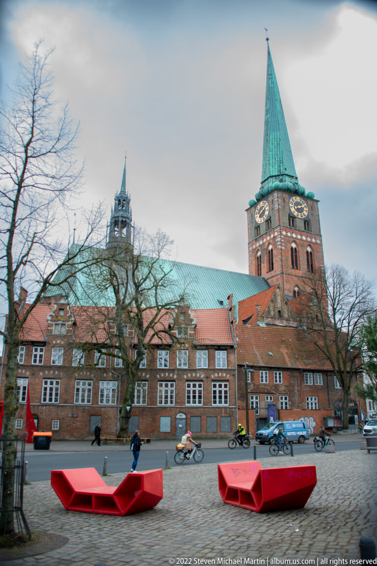 St. Jacob's Church tower and long brick roof behind historic buildings and bright red benches in an open square.