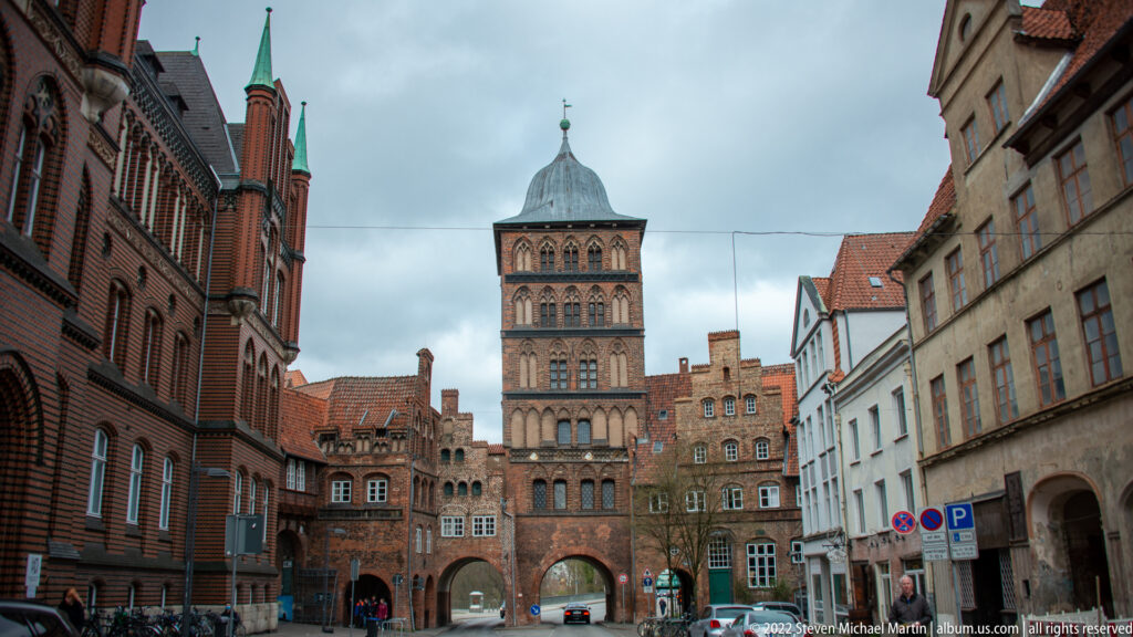 Medieval brick gateway with arched passage, tall square tower, and adjoining historic buildings along a narrow street.
