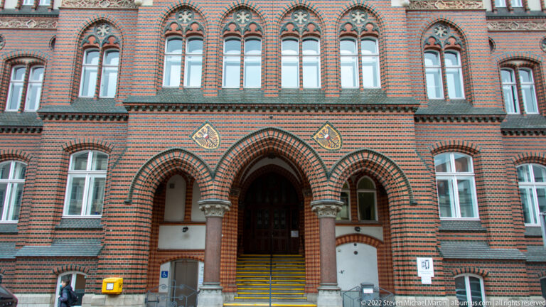 Triple pointed-arch entrance in striped red-and-dark brick with carved capitals and tall Gothic windows above.