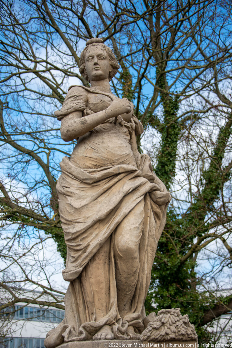 Seated stone female figure in flowing drapery holding a long narrow object beneath bare tree branches.