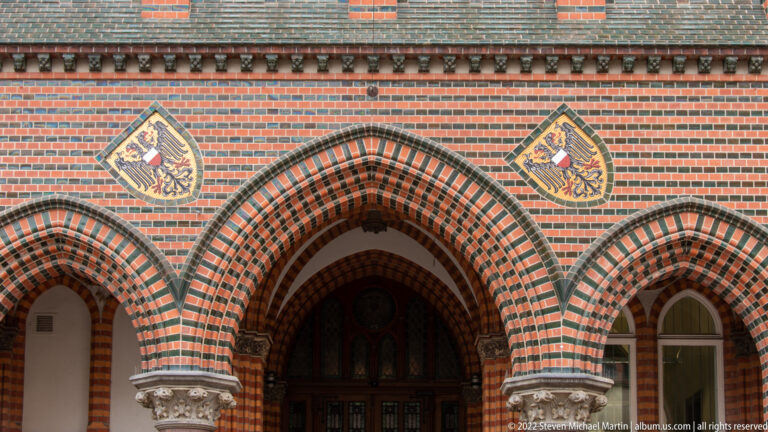 Two heraldic shields above a central pointed arch in striped red-and-dark brick masonry.