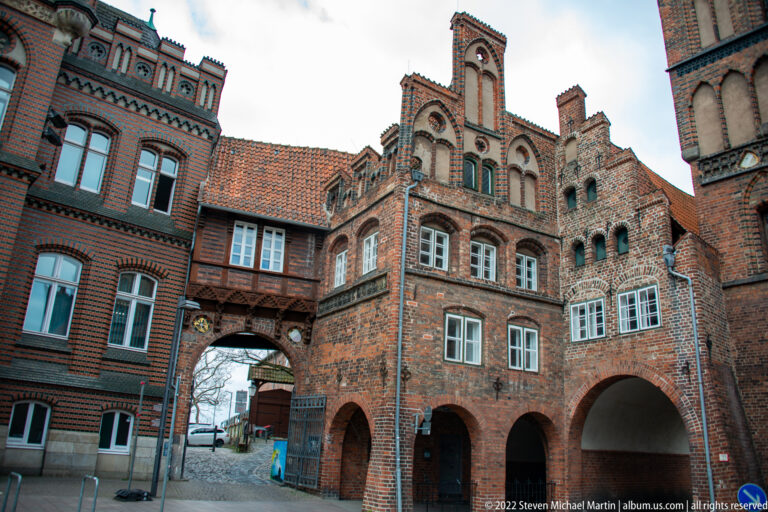 Historic brick courtyard with ornate gables, arched openings, and an enclosed bridge linking two buildings.