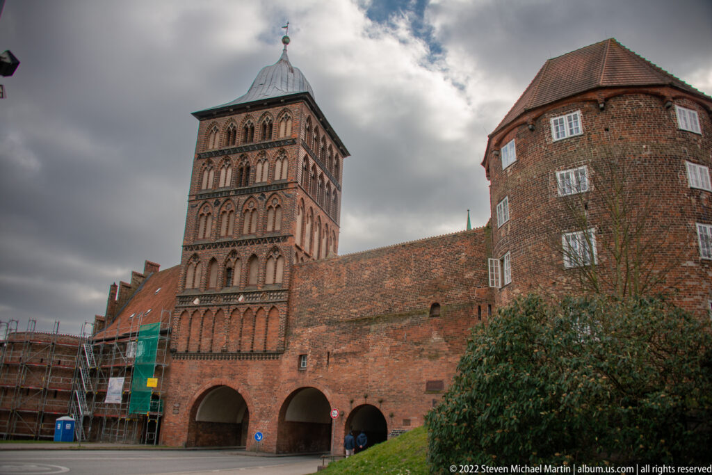Historic brick complex with tall square arcaded tower, broad wall with two arches, and rounded corner building.