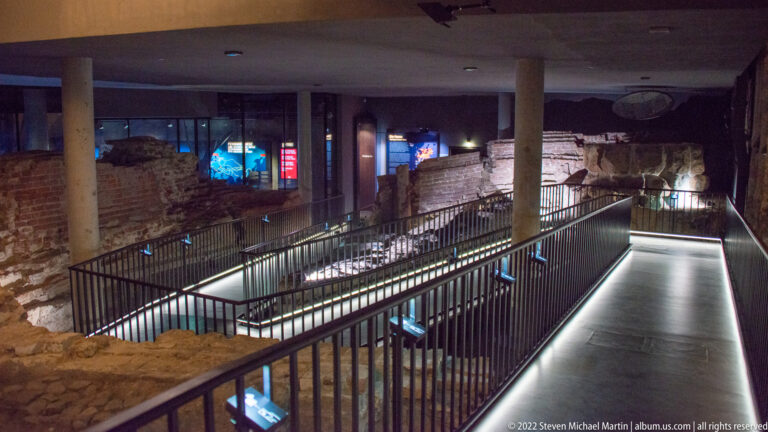 Excavated stone and brick foundations beneath a modern museum interior with black railings and illuminated walkways.