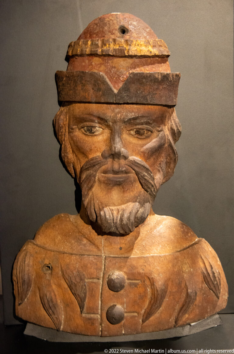 Carved wooden male bust with cap, moustache and beard, broad shoulders, and a worn brown surface.