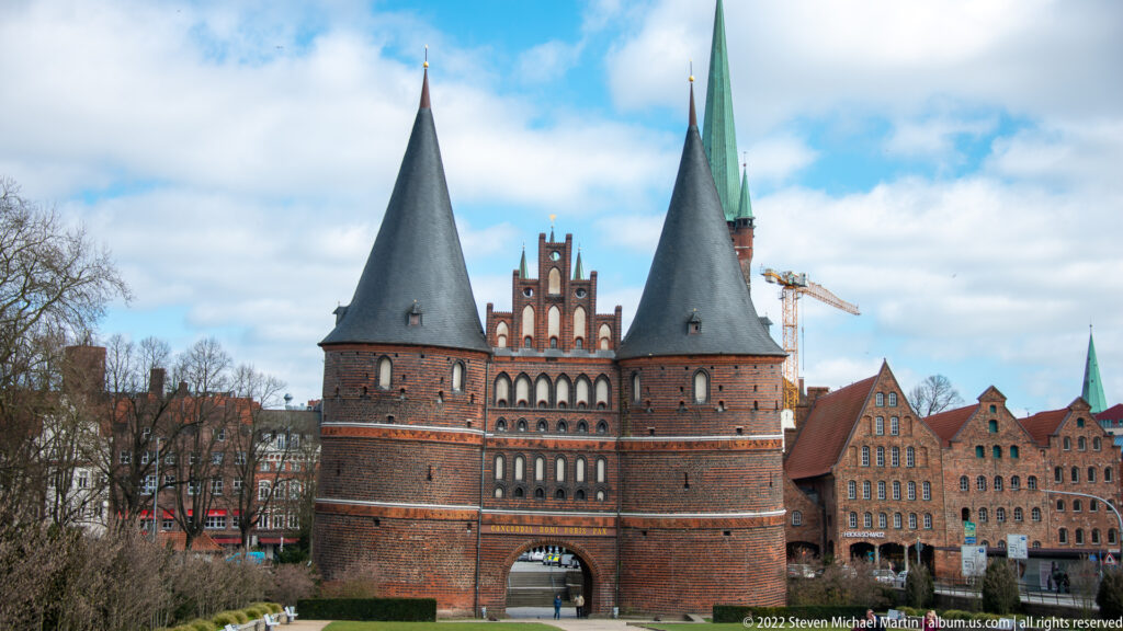 Holstentor with two round brick towers, conical roofs, central stepped gable, arched gateway, and historic buildings nearby.