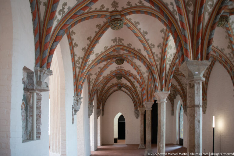 Long Gothic passage with richly painted rib vaults in red, green, and ochre above pale walls and brick flooring.
