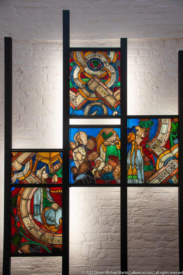Colorful figural stained-glass fragments mounted in a black grid with broad blank spaces between surviving sections.