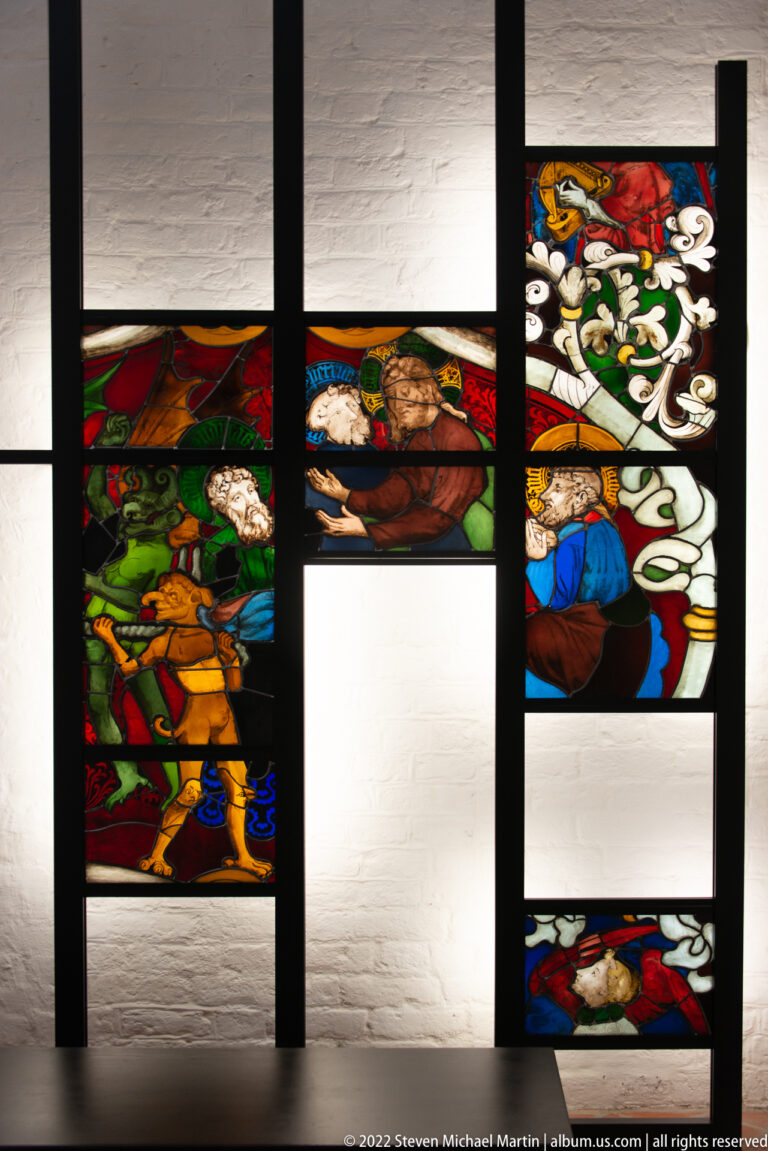 Historic stained-glass fragments with figures and ornamental motifs displayed in a black frame against a white wall.