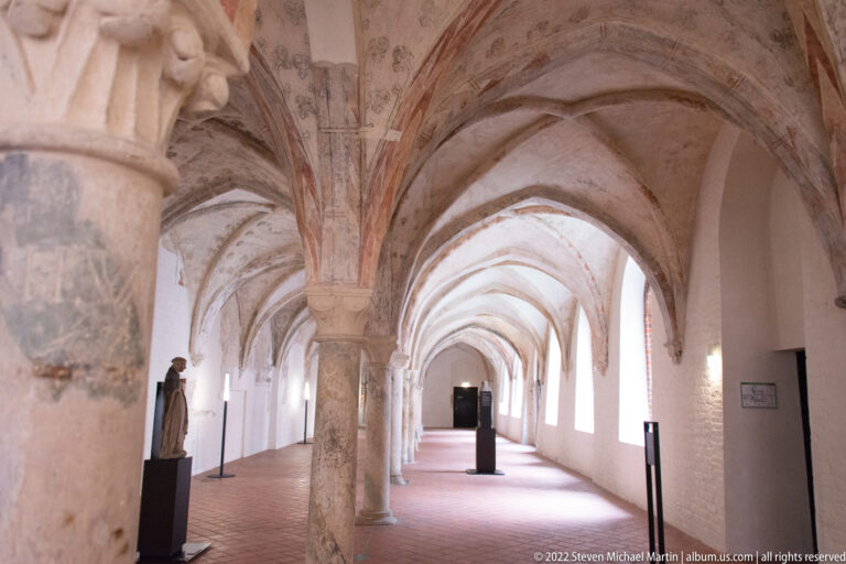 Long vaulted historic passage with pointed arches, heavy columns, brick floor, and daylight entering along one side.
