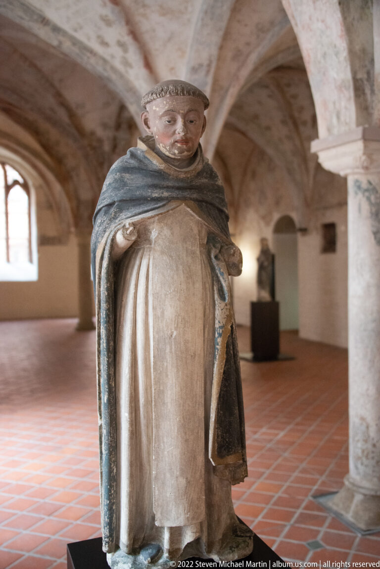 Standing painted religious figure in pale robe and blue-gray mantle displayed on a dark pedestal in a vaulted room.