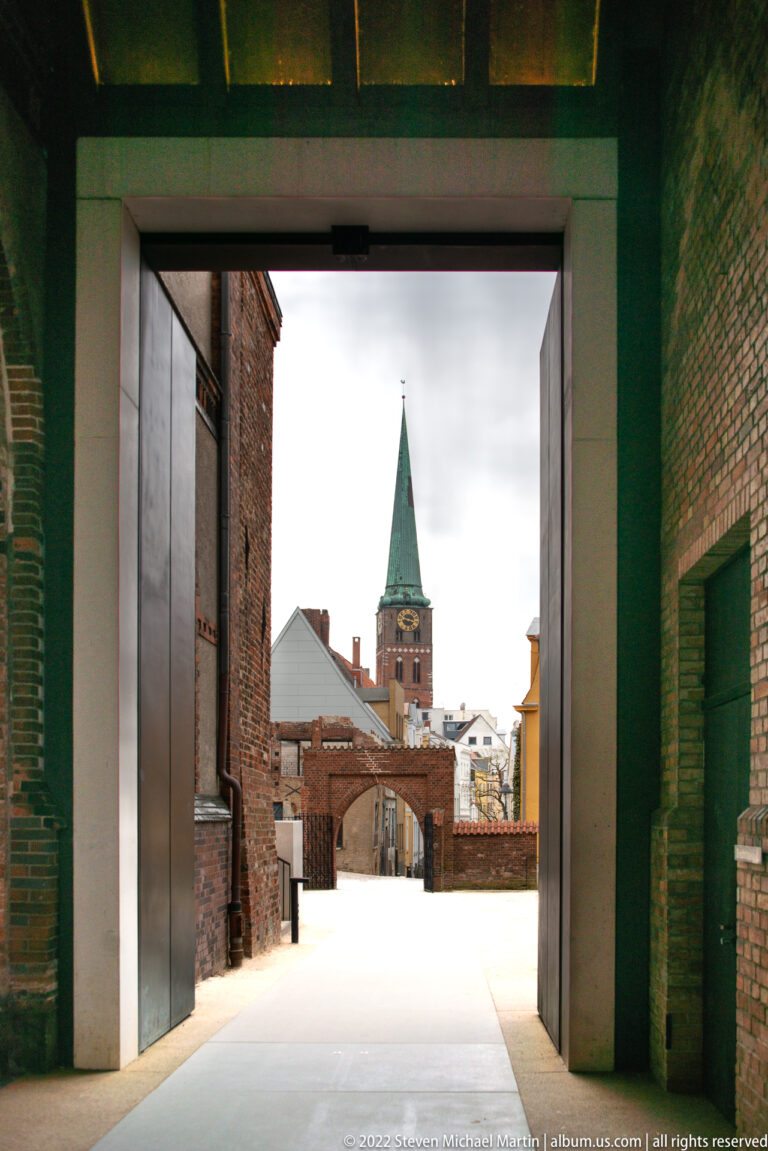 Large dark passage framing a courtyard and distant green-spired brick church tower.