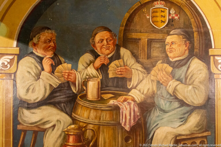 Painted scene of three men in historical dress drinking around a barrel beneath an arched doorway with heraldic symbols.