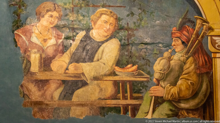 Fragmentary historic wall painting with two figures at a table and an armed figure standing at right.