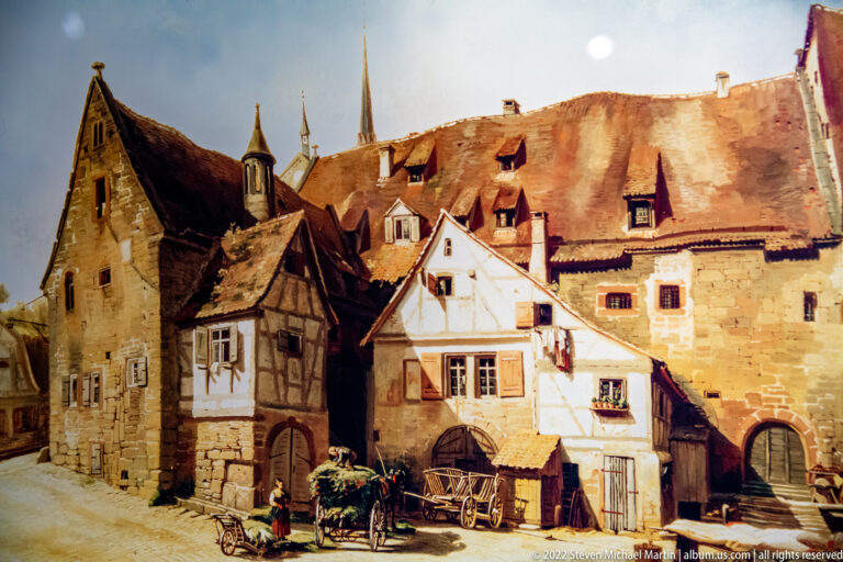 Color historical illustration of stone and half-timbered buildings, wagons, and figures beneath church spires.