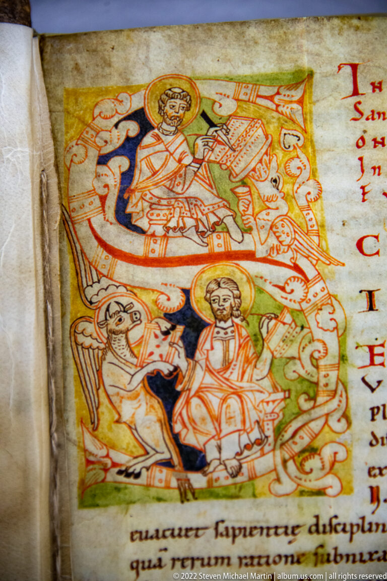 Colorful medieval manuscript illumination with a central sacred figure, surrounding figures, decorative forms, and red text on parchment.