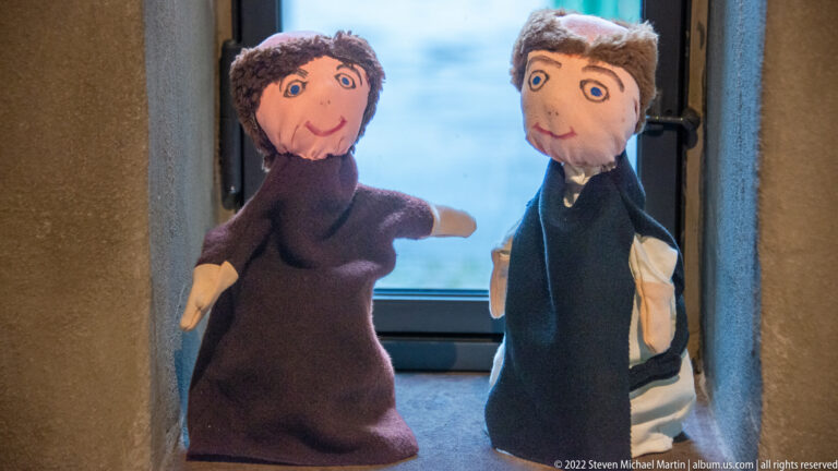Two fabric hand puppets with oversized faces stand side by side on a ledge beside a window.