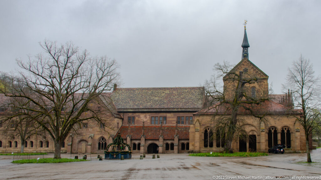 Arcaded sandstone monastery range with a central gabled pavilion, slender turret, and fountain in the courtyard.