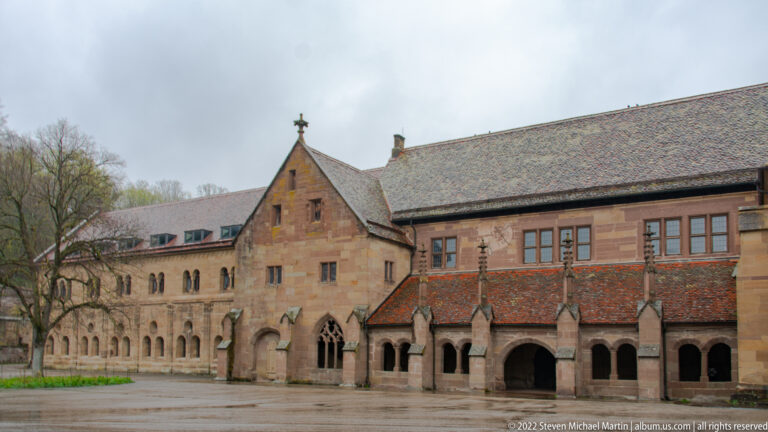 Long stone monastery range with ground-floor arcades, upper windows, projecting gable, and tiled roof beside the courtyard.