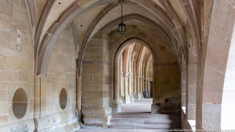 Stone cloister passage with ribbed vaults, pointed arches, a hanging lantern, and an adjoining arcade visible through the end arch.