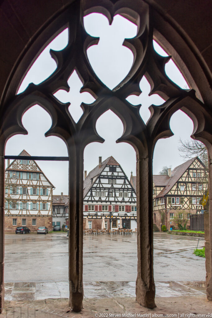 Three Gothic tracery openings frame a paved courtyard and half-timbered buildings beyond.