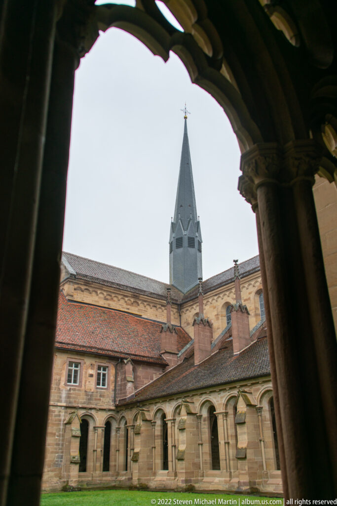 Dark cloister arch framing a tiled courtyard range and a slender church tower.