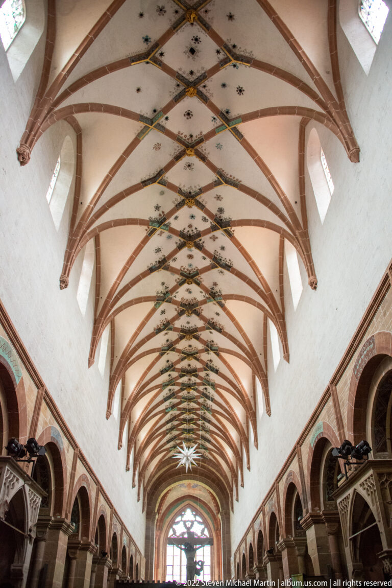 Upward view along a church nave with red rib vaults, pale plaster, and small painted ceiling motifs.