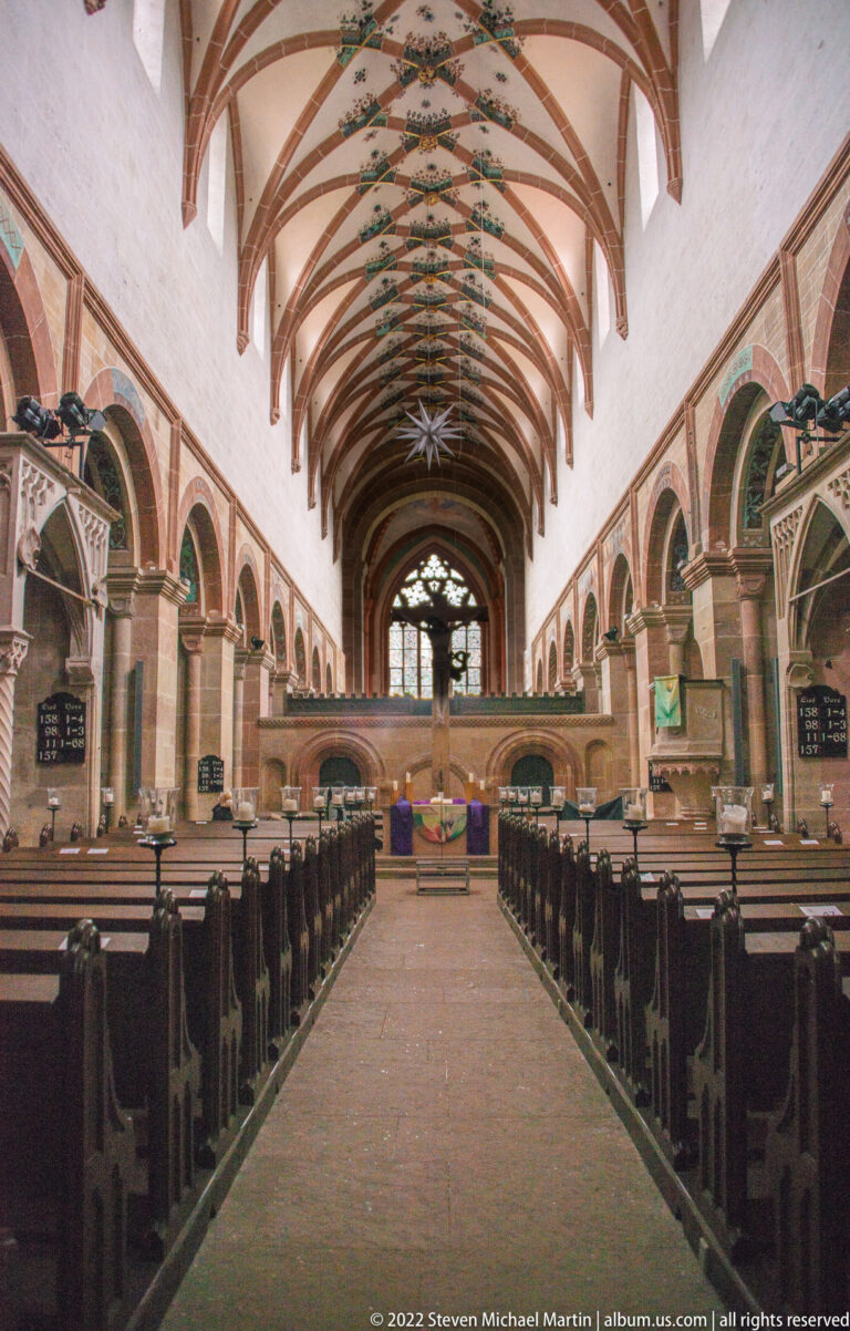 Central aisle of a monastic church with dark pews, pointed arcades, and painted rib vaults.