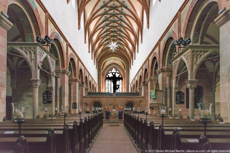 Closer axial view of the church nave with pews, side furnishings, pointed arcades, and painted rib vaults.