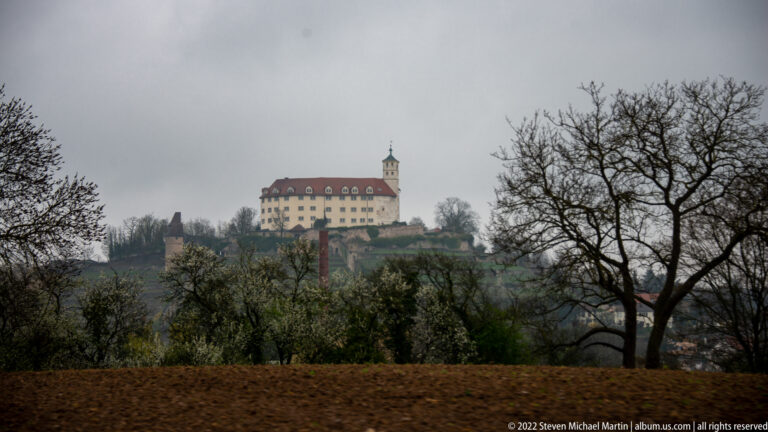 Large pale historic building with a red roof and small tower standing on a ridge beyond fields and bare trees.