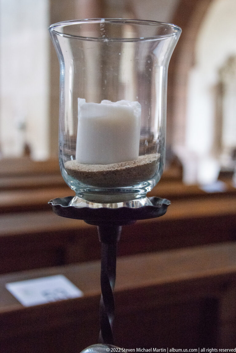 White candle in a clear glass holder on a dark metal stand, with wooden pews blurred behind.