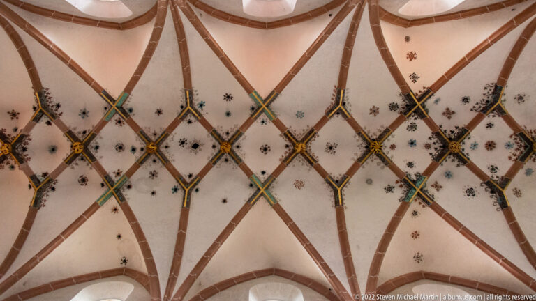 Close upward view of intersecting red ribs, colored bosses, and small painted motifs on a pale church vault.