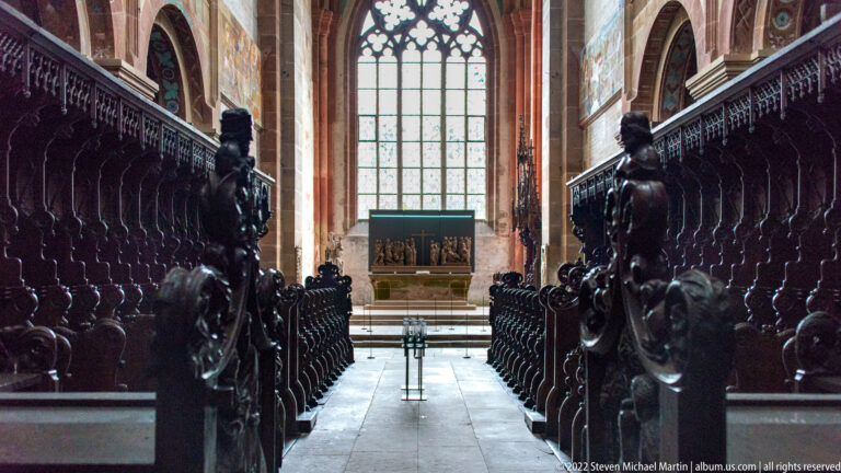 Dark carved choir stalls on both sides of a narrow aisle leading toward a tall window and altar.