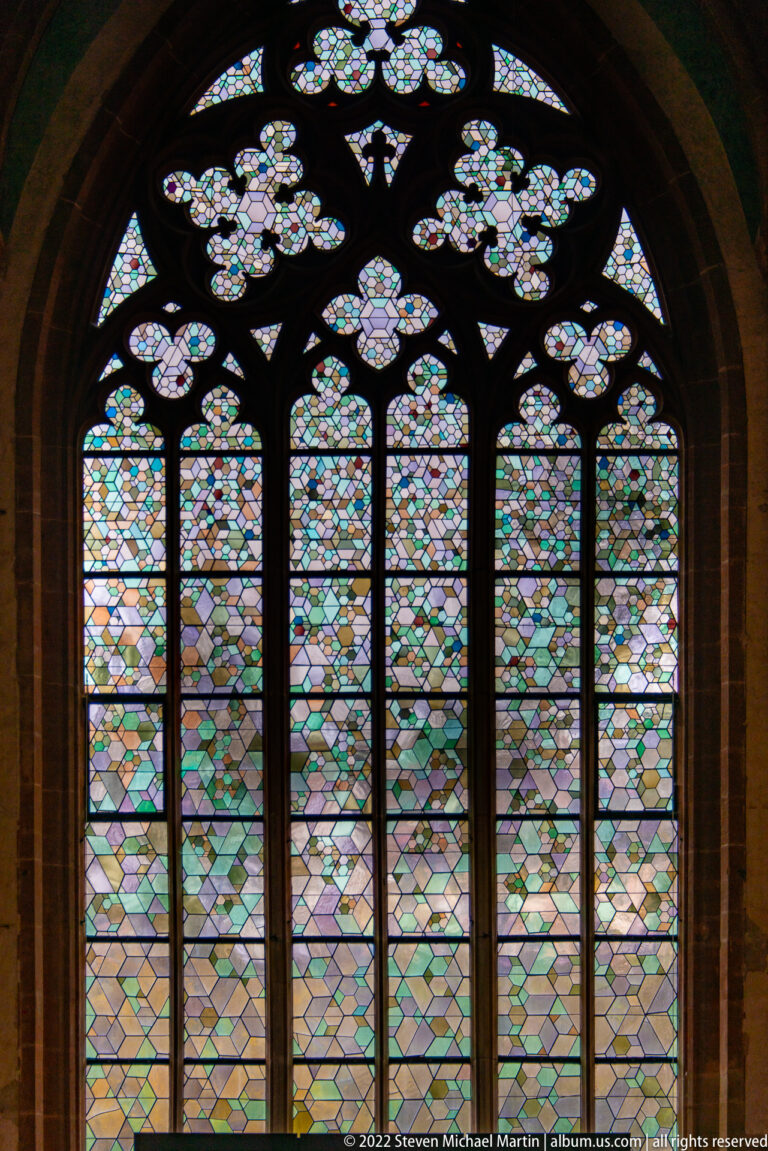 Large Gothic tracery window with tall lights filled by patterned pastel stained glass.