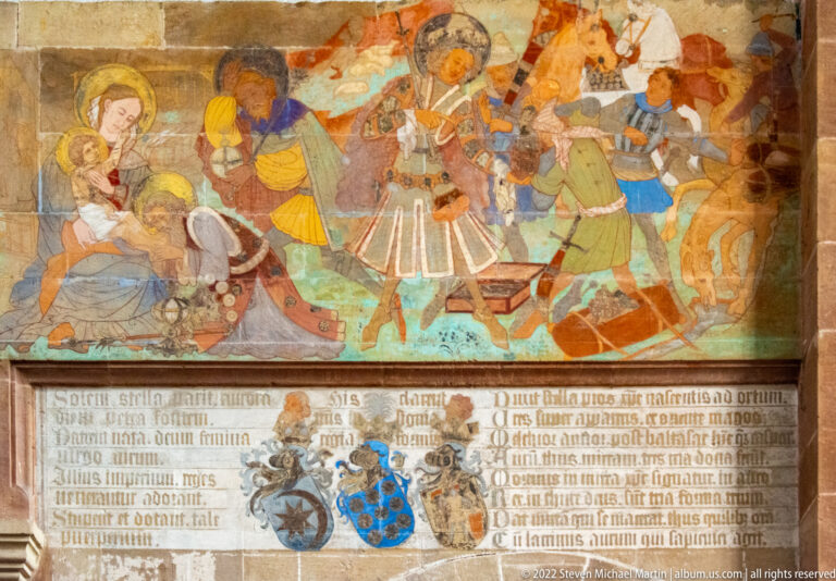 Narrative wall painting with colorful figures above a long inscription and several heraldic shields.