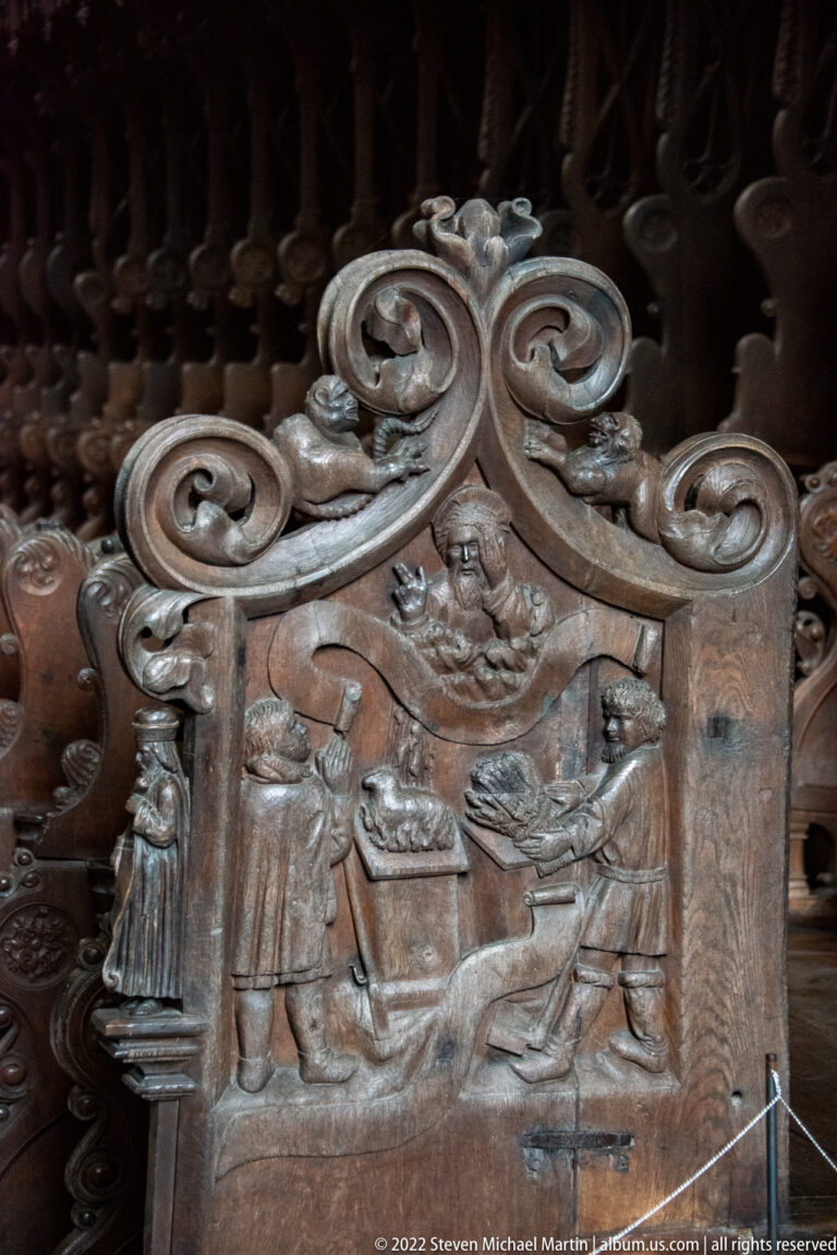 Close carved wooden choir-stall panel with scrolling ornament and a small relief scene of figures.