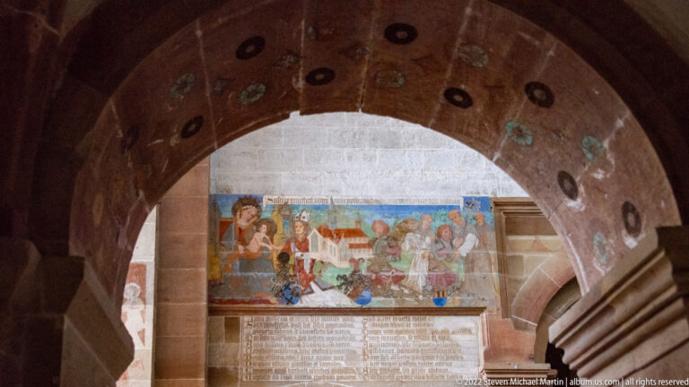 Painted archway framing a narrative mural and inscription panel, with a staircase at right.