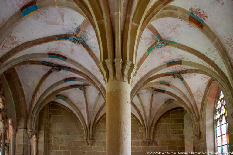 Central stone column supporting branching rib vaults with faded painted decoration on the ceiling.
