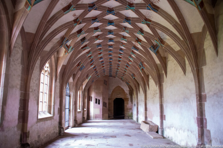 Long empty vaulted room with tall side windows, painted ribs, and a dark pointed doorway at the far end.
