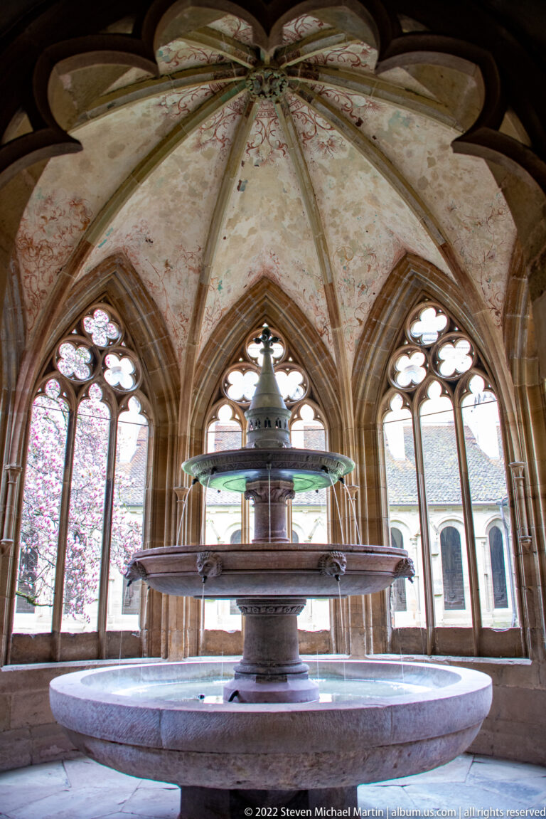 Tiered stone fountain in a polygonal vaulted room surrounded by pointed Gothic windows.