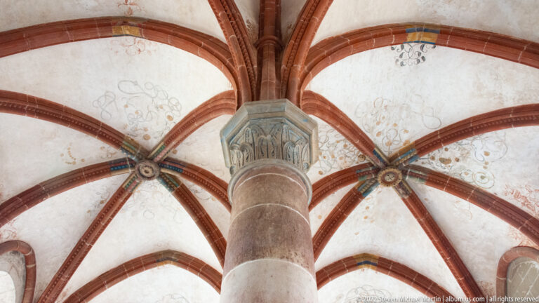 Stone column with carved capital supporting radiating red ribs beneath a lightly painted vault.