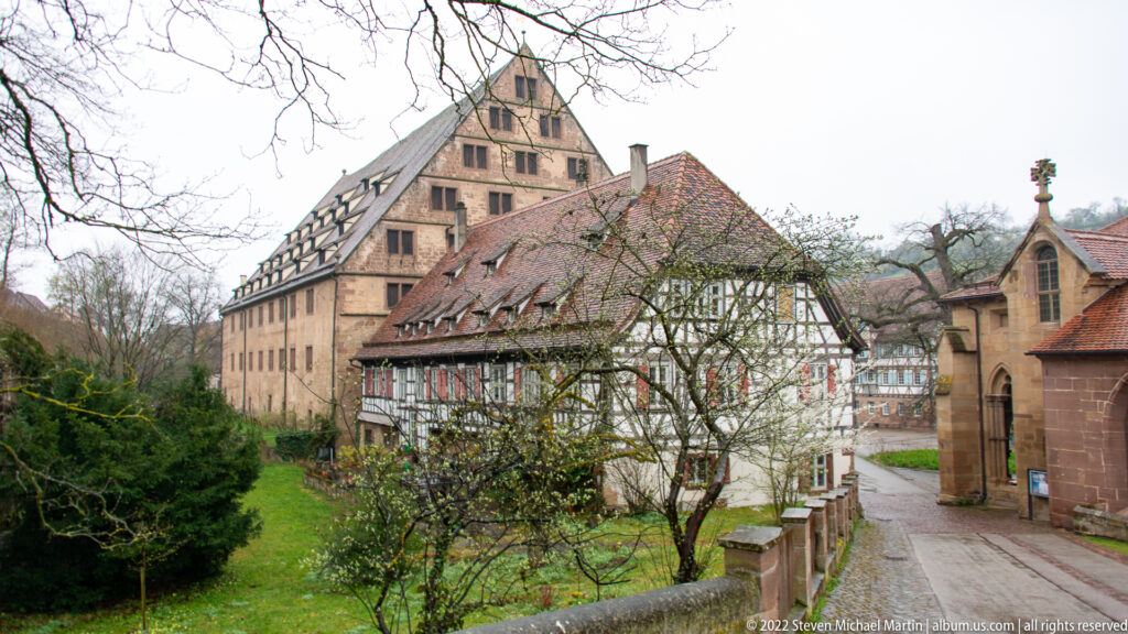 Half-timbered house beside larger stone buildings and red tile roofs along a narrow lane in Maulbron.