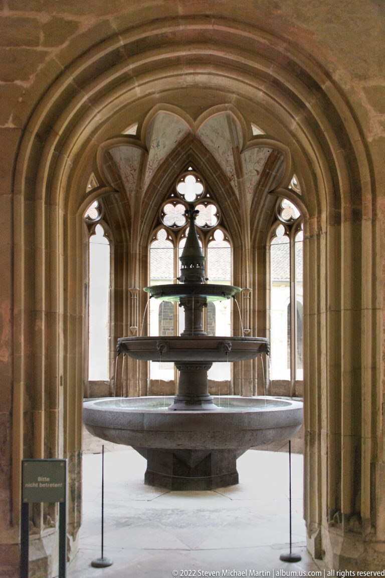 Tiered stone fountain centered beneath deep Gothic arches and traceried windows in the monastery fountain house.